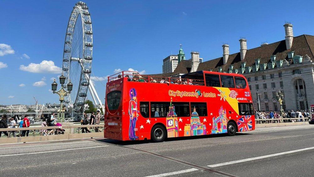Red hop-on hop-off sightseeing bus driving past the London Eye and historic buildings in London