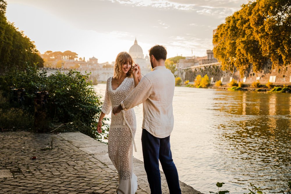 A man twirling a woman by a river surrounded by small trees with a chapel behind them.