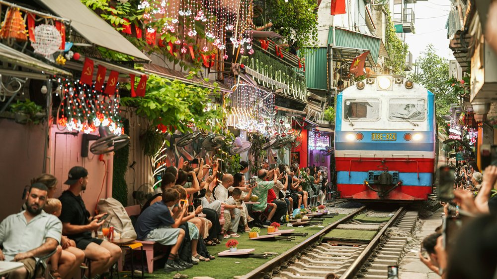 A train passing through an alleyway full of people sitting down and storefronts.