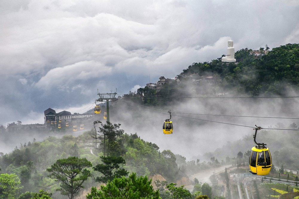 Yellow cable cars moving across a cloudy mountaintop covered with greenery.