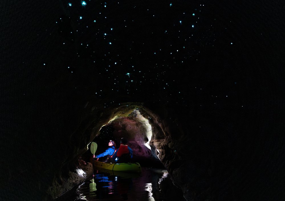 A dark cave with lit up spots all over and a small entrance that a 2-person kayak is passing through.