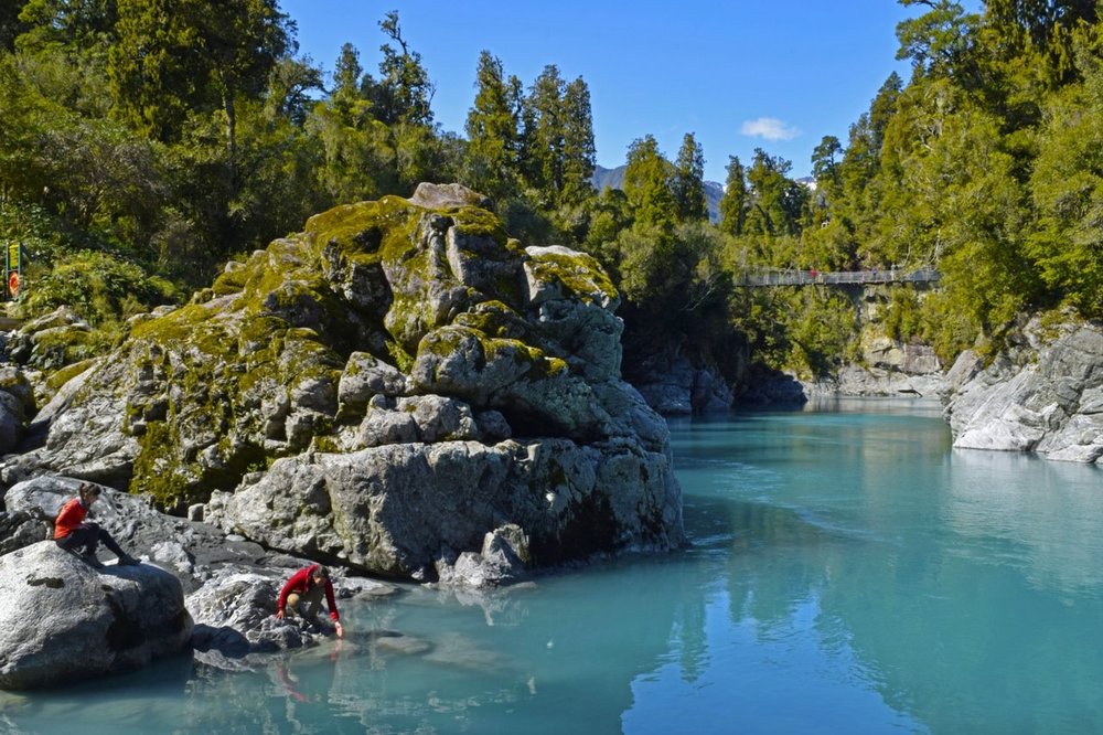 A large rock formation with people next to it, in beside tall green trees and a turquoise body of water
