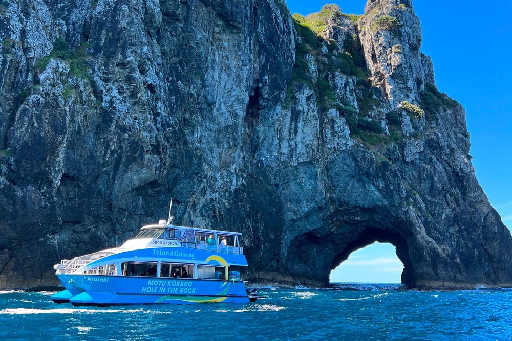 A boat in front of a large rock formation with a large hole on its right side.