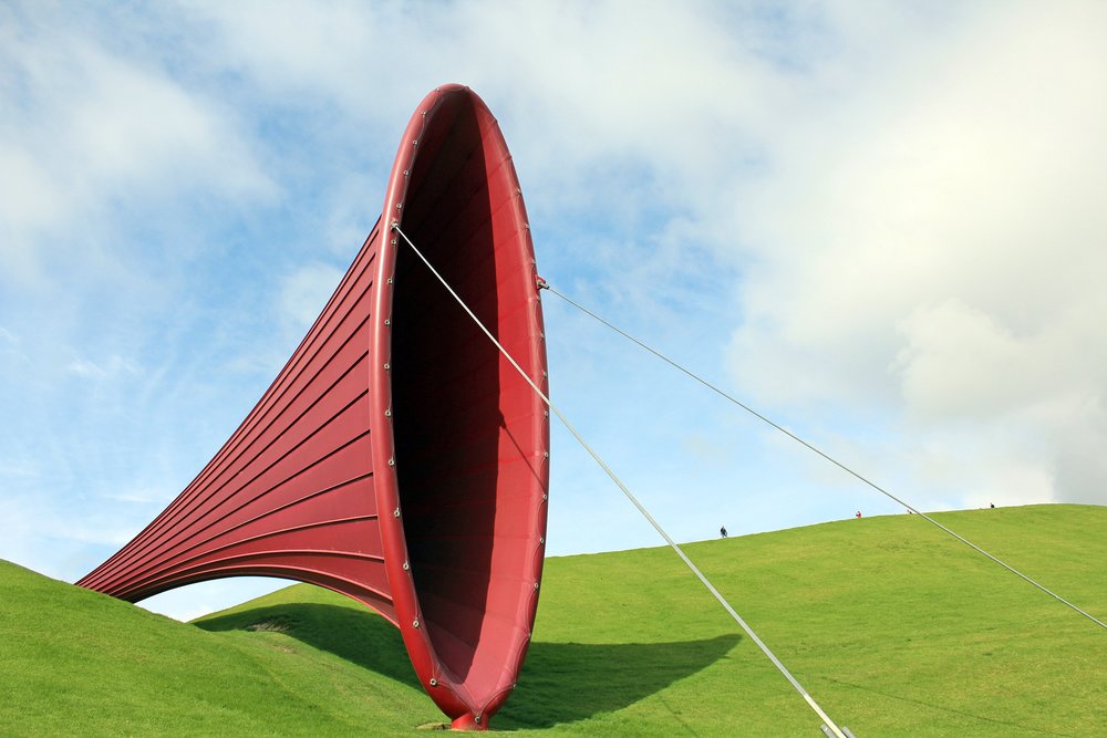 A large red cone sculpture on a green hill against a blue, cloudy sky.