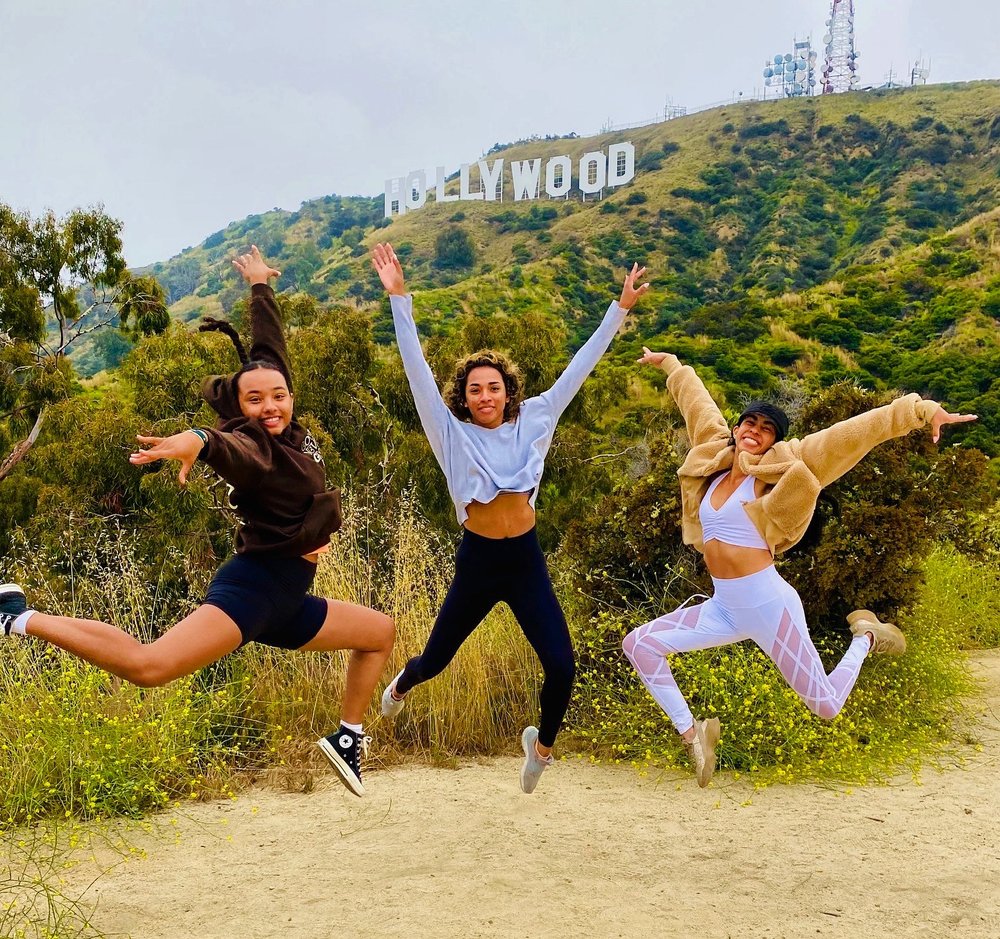 Three people jumping mid-air on a trail with the Hollywood Sign visible behind them on the hill