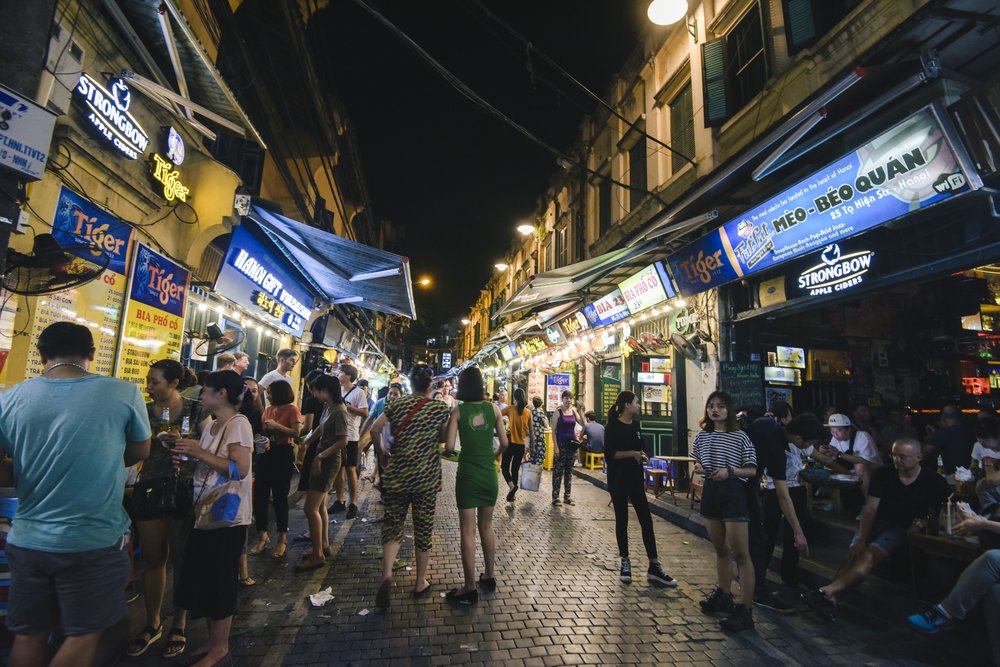 A busy street scene in Hanoi's Old Quarter with dozens of motorbikes, street vendors, and traditional buildings.