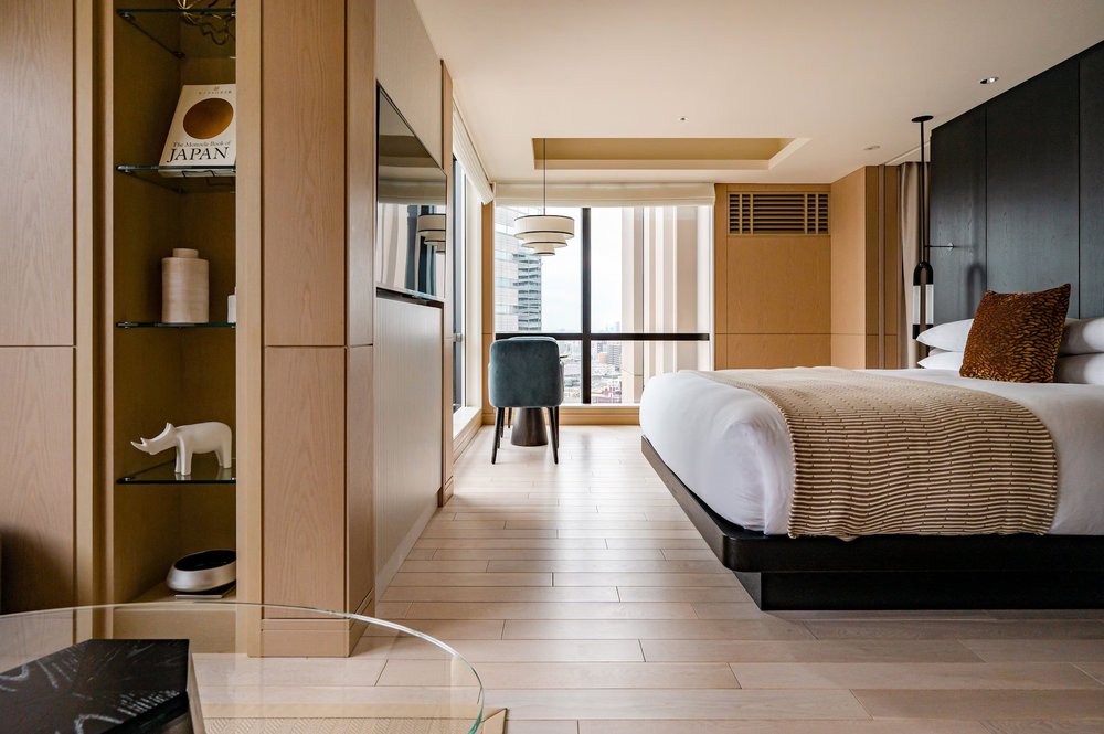 A hotel room showing a bed, a coffee table, a chair, a shelf, and a floor-to-ceiling window