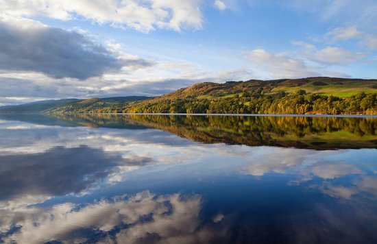 Autumn trees reflecting on the still water of Loch Ness under blue skies.