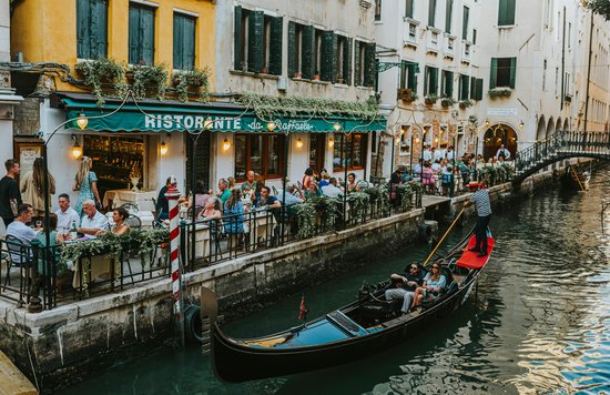 Outdoor restaurants along a Venice canal with diners and a gondola passing by