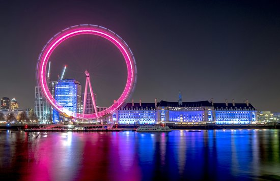 London Eye at night illuminated in pink with city lights reflecting on the Thames