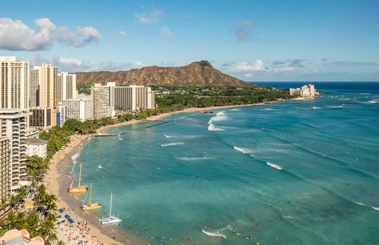 Tall buildings beside a wide stretch of sand and a large body of water, with a volcano behind it.