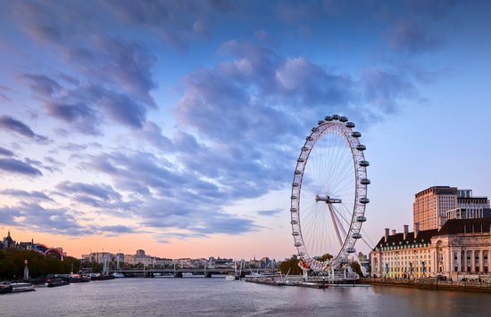 The London Eye beside the River Thames at sunset, with city buildings and a cloudy blue sky in the background.