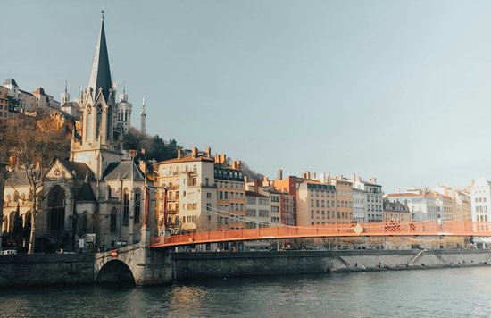 Side-by-side mid-rise buildings and a cathedral above a river with an orange bridge stretching out of frame. 