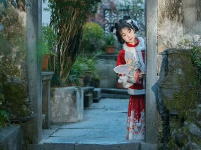 红底白纹绣衣女童,轻扶石框探身而出,手持蝶形团扇,青石板巷与绿植相映,尽显徽州庭院里的灵动与天真