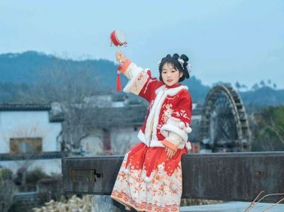 红底白纹绣袍女童,手持拨浪鼓坐于石栏,背景是白墙黛瓦与大水车,远山含黛,尽显徽州冬日里的鲜活与喜庆