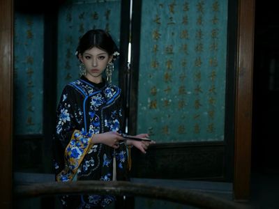 黑底蓝花清式绣衣女子,手持折扇立于青底金书屏前,银饰垂坠,眉眼沉静,尽显徽派古宅中大家闺秀的典雅与故事感