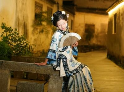 青花绣衣少女,斜倚石栏轻摇折扇,暖黄灯光映着斑驳古墙,衣袂翩跹,尽显徽州夜巷里的温婉与诗意