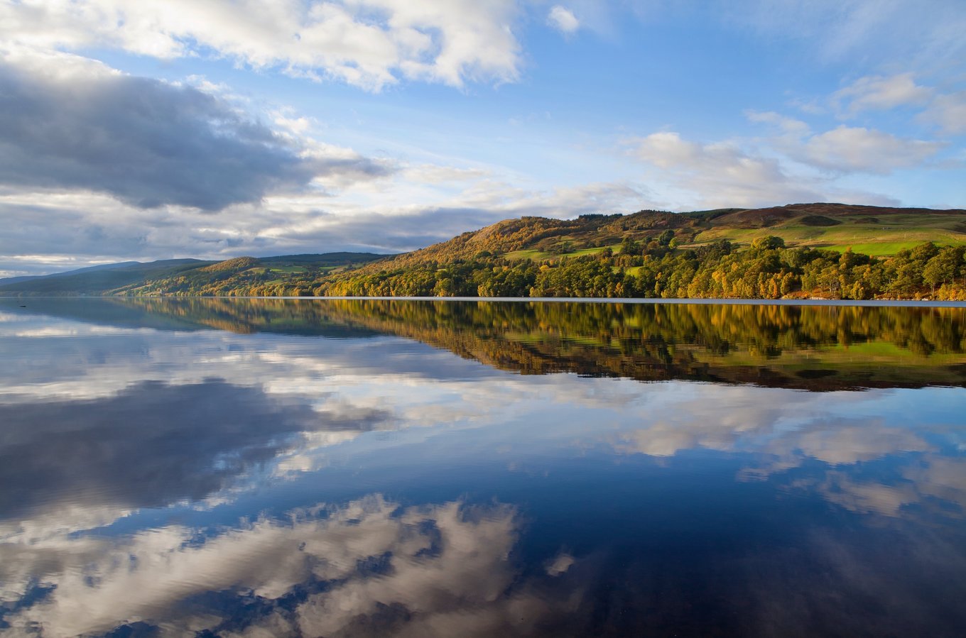 Autumn trees reflecting on the still water of Loch Ness under blue skies.