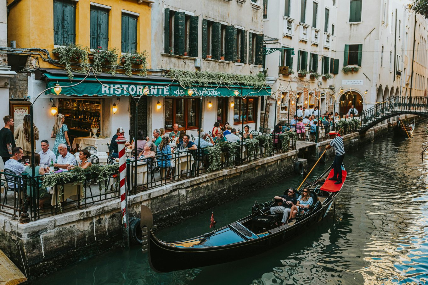 Outdoor restaurants along a Venice canal with diners and a gondola passing by