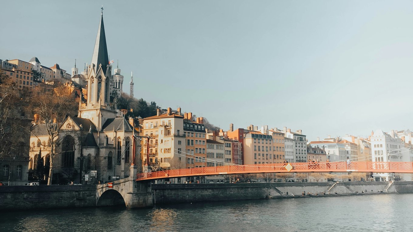 Side-by-side mid-rise buildings and a cathedral above a river with an orange bridge stretching out of frame.