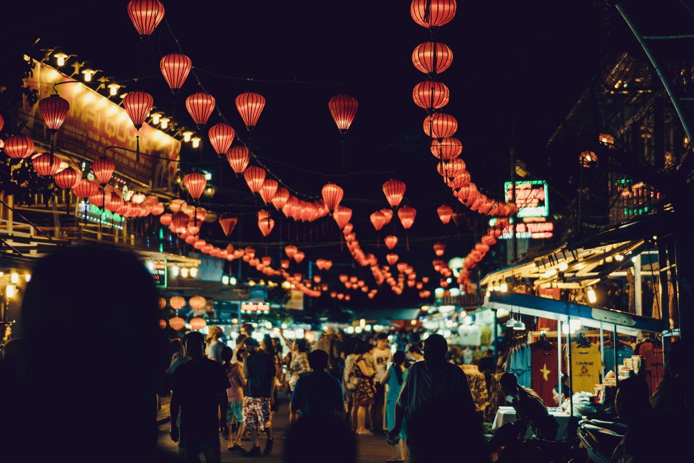 A shopping street with lamps strung across the area, lit up storefronts, and people walking.