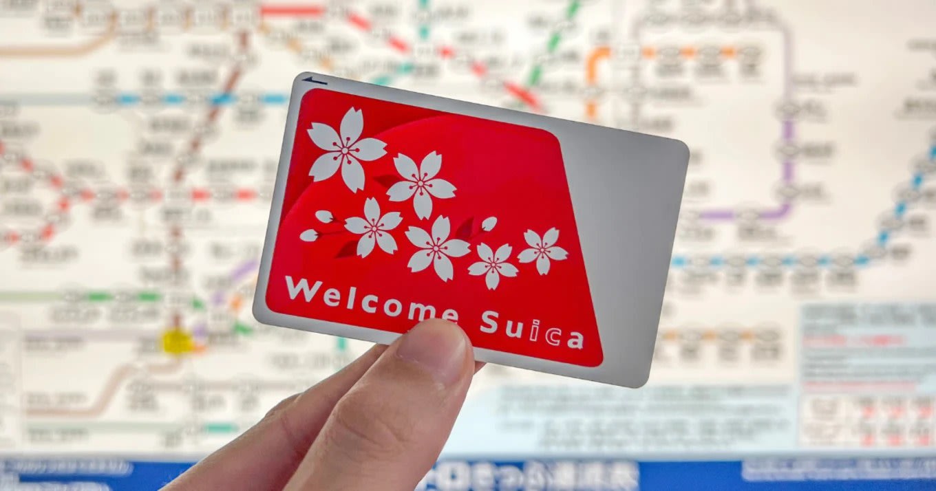 Close-up of a hand holding a red Welcome Suica IC card with cherry blossoms, train map blurred in background