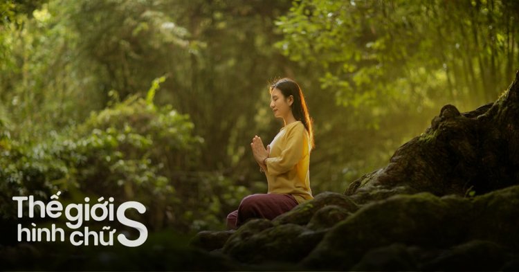A woman dressed in yellow and maroon is sitting cross-legged on rocks in a lush, green forest, practicing meditation or mindfulness.