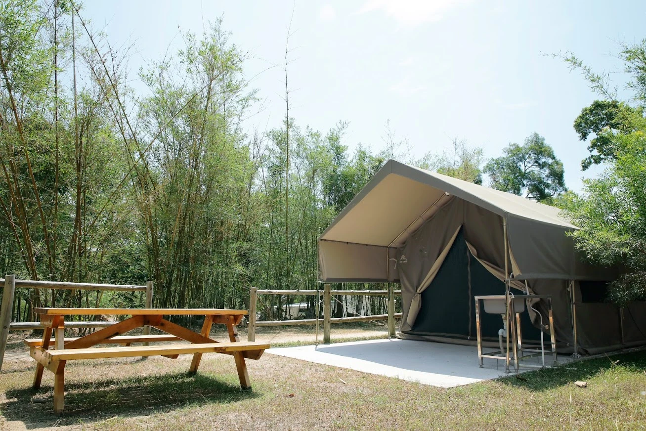 在竹林旁邊搭建的戶外帳篷，前面有一張大型木製野餐桌及遮陽傘，周圍以木圍欄分隔。