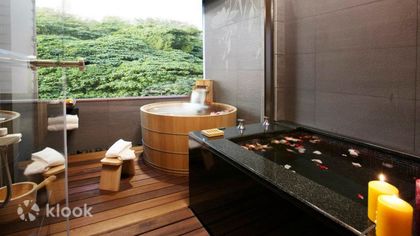 A private hot spring room with a cypress barrel tub and a black stone tub, candles lit, green hillside through the window
