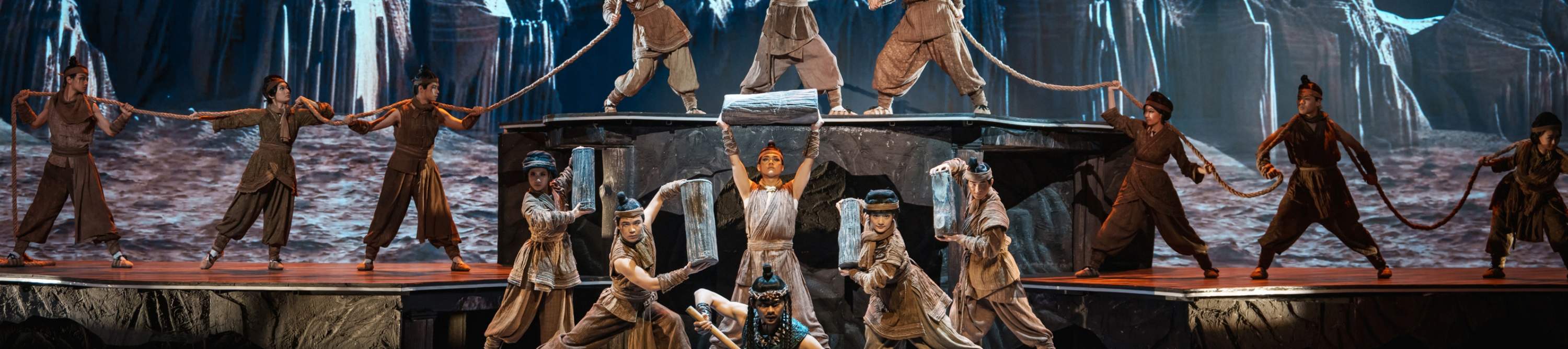 On a multi-layered stage, unified costumes, coordinated with ropes and props, and a realistic mountain rock background create a scene where their movements are orderly and full of power, as if they are performing a collective labor of an ancient tribe.