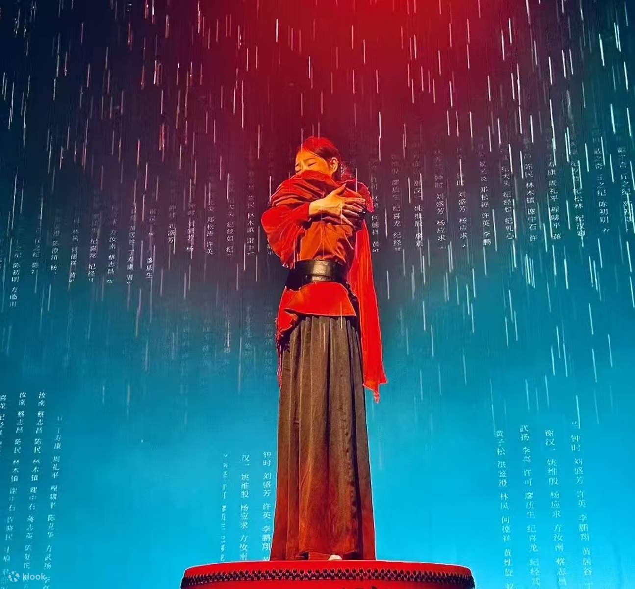 Ein rot gekleideter Schauspieler steht mit verschränkten Armen auf einer Trommel, ein Regenvorhang im Hintergrund unterstreicht die Namen und Texte, und rote und blaue Lichter erzeugen ein Gefühl von Geschichte.