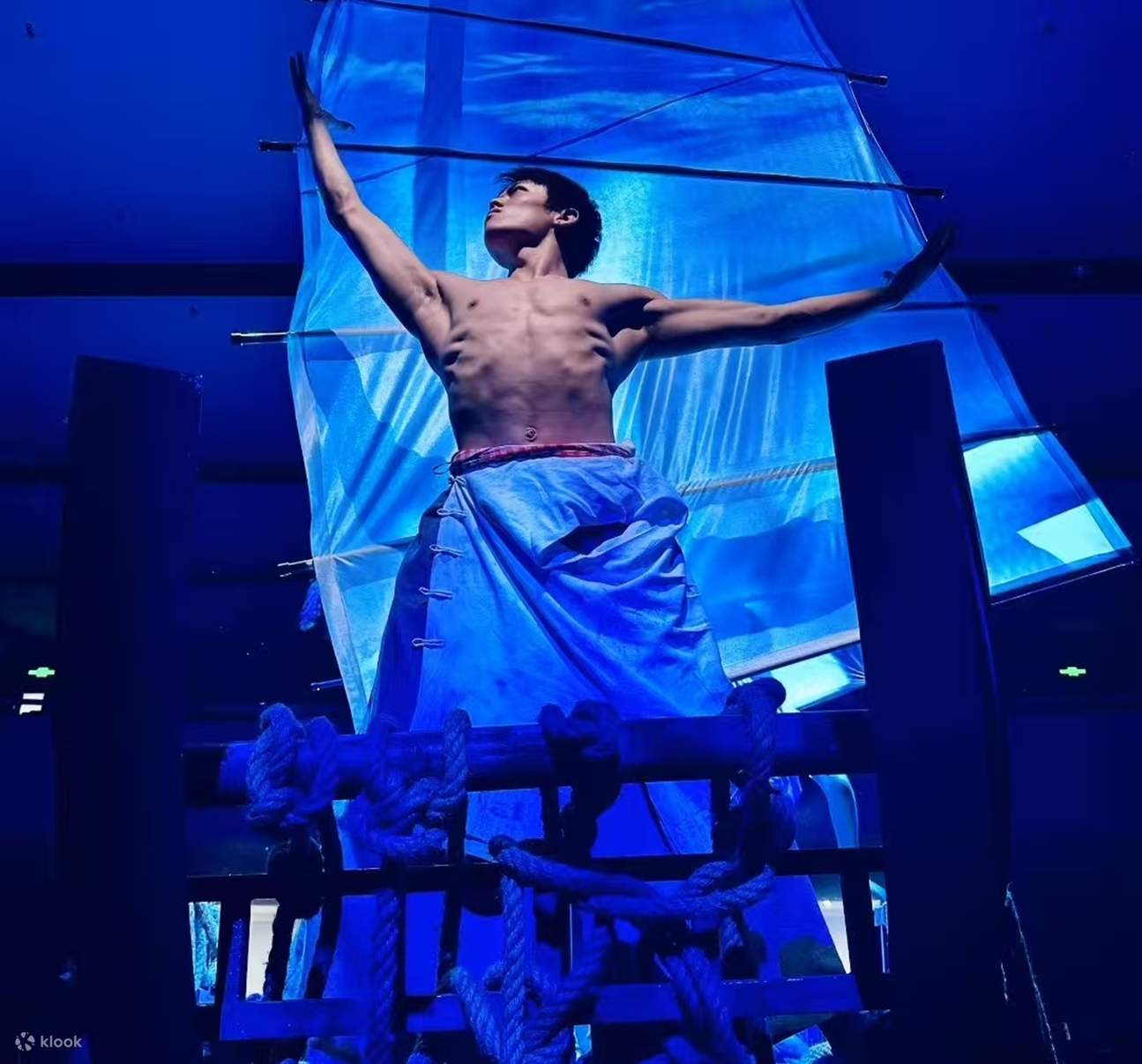 El actor, con el torso desnudo y una falda de tela azul, se alza sobre un atrezo con forma de barco y extiende los brazos. La iluminación azul resalta el ambiente escénico de un pueblo de pescadores, creando una atmósfera llena de tensión.