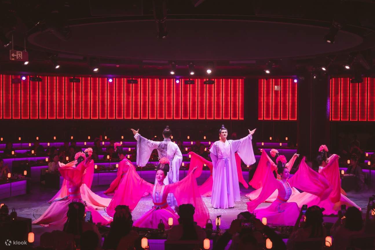 Para penari dengan kostum tari tradisional berwarna ungu muda bergerak dengan anggun, gerakan mereka halus dan indah, dengan koordinasi yang sempurna. Para penampil pria dengan jubah putih berdiri di samping.