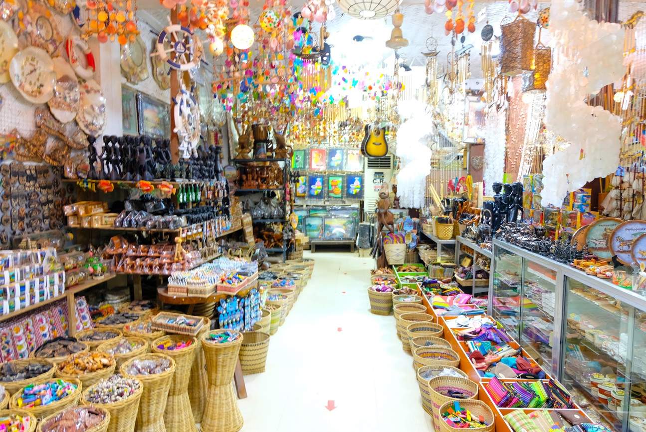 纪念品商店的内部，物品排列在单通道的左右两侧。左侧是微型雕塑，小纪念品篮子，可以看到风铃悬挂在天花板和墙壁上