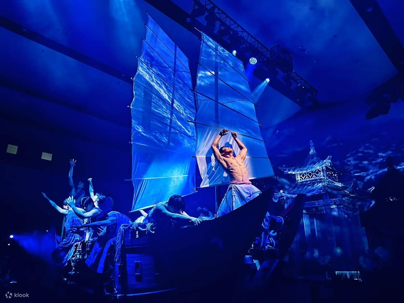 На реквизите в виде лодки актеры синхронно вытягивают руки, синие паруса переплетаются со светотенью водных узоров, воссоздавая сцену рыбацкого труда.