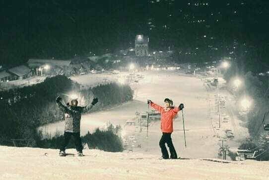 Dua pemain ski berdiri di lereng bersalju, siap untuk meluncur turun