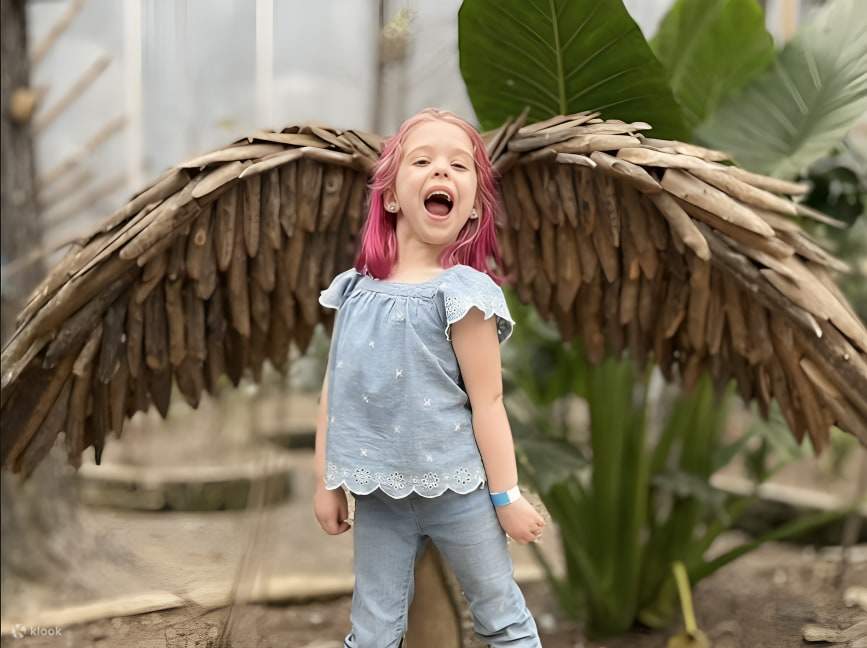 Una niña posa alegremente para una foto frente a un conjunto de alas falsas y vibrantes, sonriendo ampliamente.