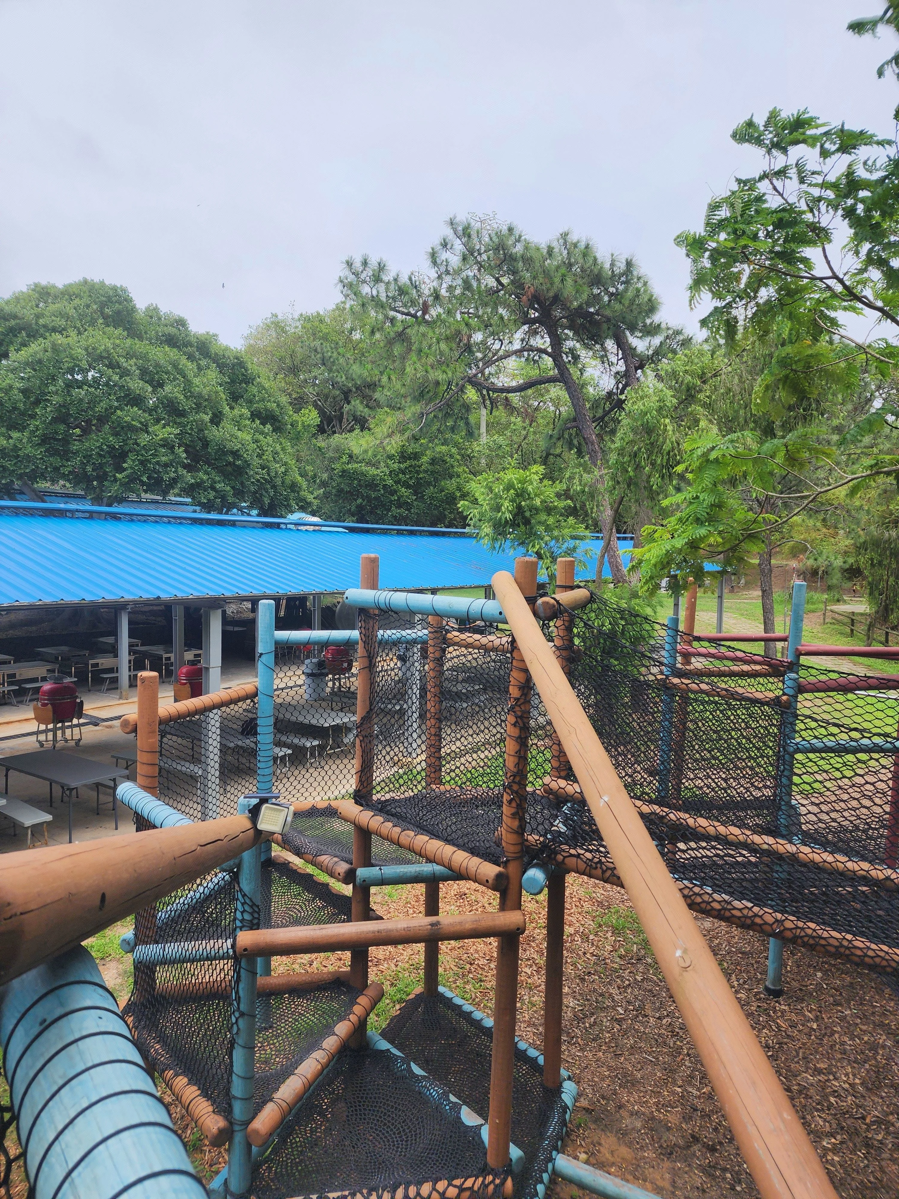 兒童戶外攀爬網遊樂場裝置，由木柱和黑色安全網組成，背景是樹林和藍色屋頂建築。